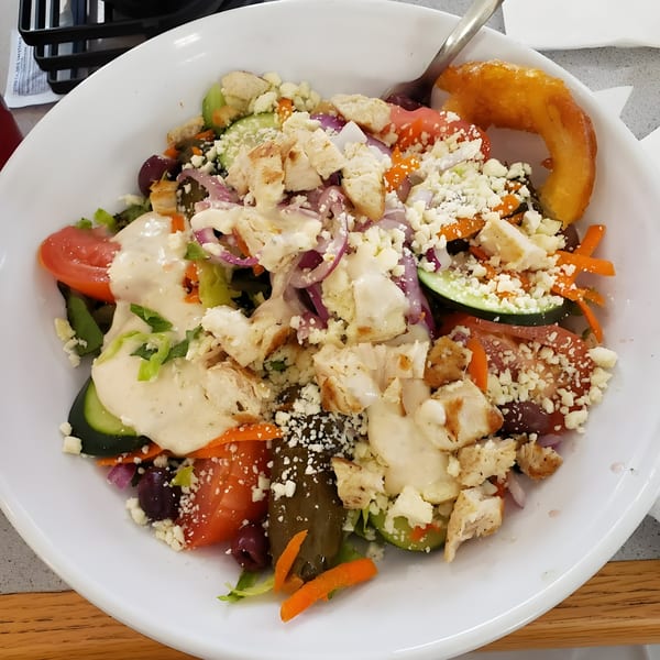A large Greek salad with grilled chicken, feta, olives, and a golden onion ring in a white bowl.